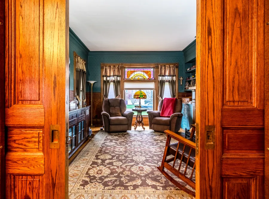 A richly furnished Victorian-style living room seen through open wood pocket doors, with a stained-glass window and patterned rug