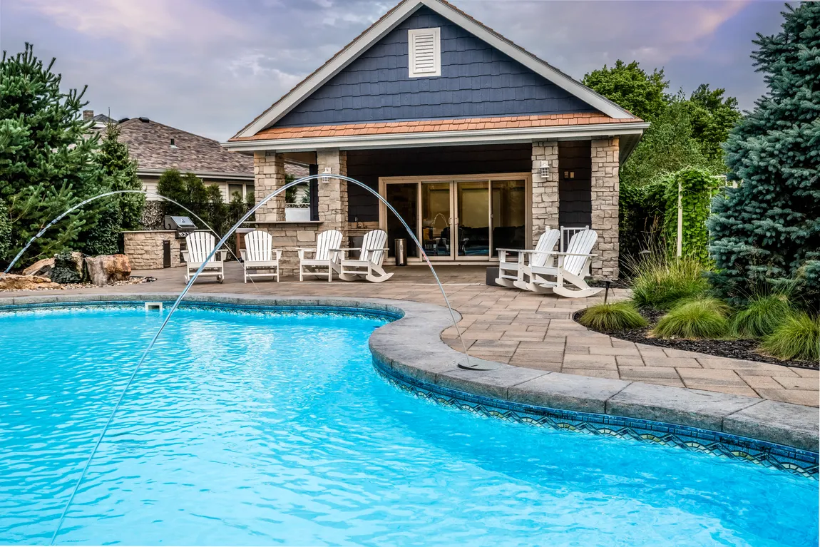 Twilight view of a stone and shingle pool house with a lit backyard pool